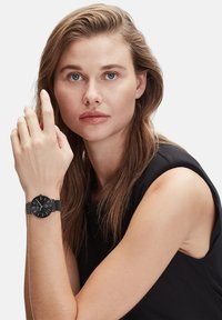 Schwarze Armbanduhr mit rundem Zifferblatt, Mesh-Armband und weißen Stundenzahlen, getragen an einer hellhäutigen Hand. Das Model hat lange Haare und trägt ein schwarzes ärmelloses Oberteil.