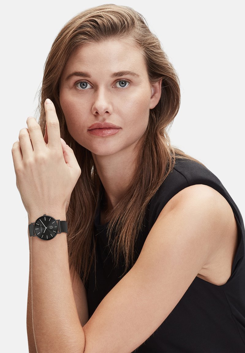 Schwarze Armbanduhr mit rundem Zifferblatt, Mesh-Armband und weißen Stundenzahlen, getragen an einer hellhäutigen Hand. Das Model hat lange Haare und trägt ein schwarzes ärmelloses Oberteil.