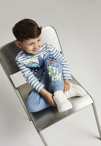 Chemise à manches longues rayée bleue avec des personnages de dessins animés ; jogging bleu assorti avec le texte "Stitch" ; baskets blanches ; pose assise sur une chaise.