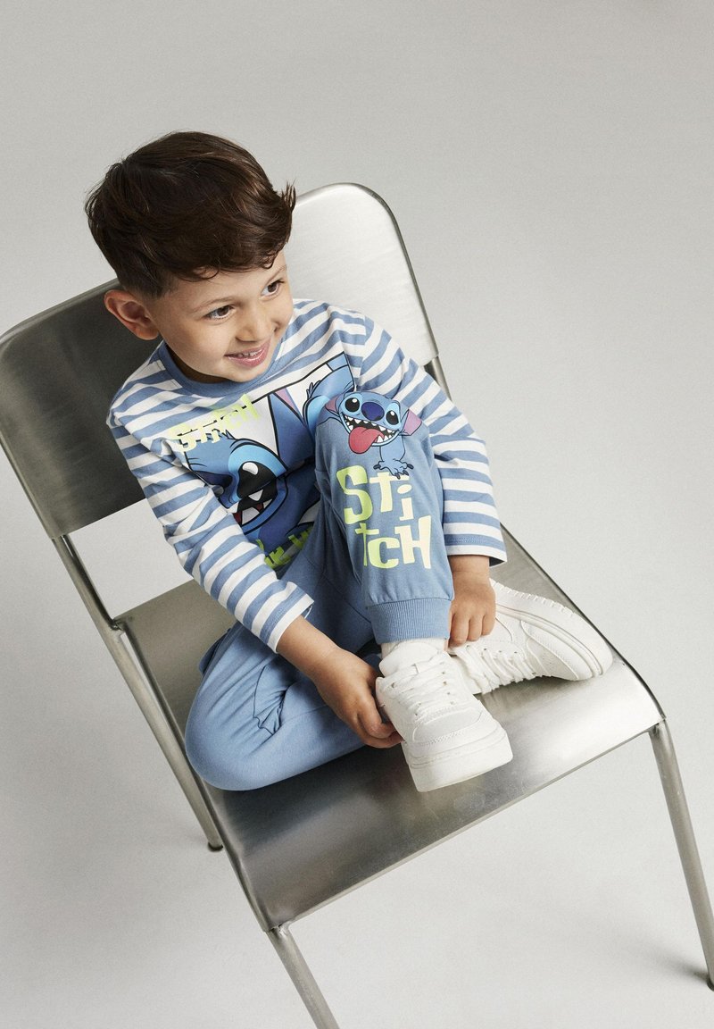 Chemise à manches longues rayée bleue avec des personnages de dessins animés ; jogging bleu assorti avec le texte "Stitch" ; baskets blanches ; pose assise sur une chaise.