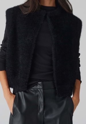 Vrouw draagt een zwart pluizig mouwloos vest over een zwart top met lange mouwen en hoog getailleerde zwarte leren broek met handen in de zakken.