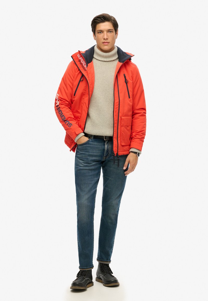 Chaqueta acolchada roja con acentos en azul marino, bolsillos con cremallera y detalles de logotipo. Combinada con un suéter de punto gris, jeans azules y zapatos negros.