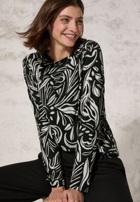 Schwarzes Langarmoberteil mit weißen Blumendetails, aus weichem Stoff, Rundhalsausschnitt, entspannte Passform, kombiniert mit schwarzen Hosen, Model sitzt.