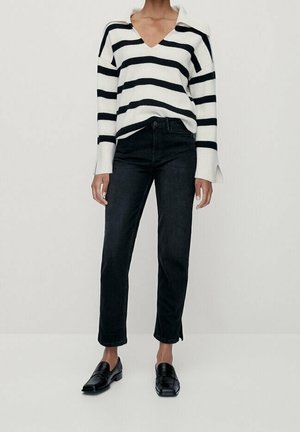 Personne portant un pull blanc avec des rayures horizontales noires, un jean noir droit avec des fentes latérales, et des mocassins noirs.