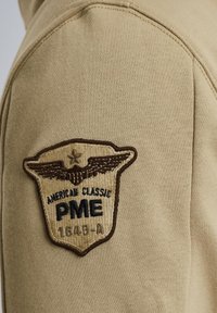 Beige Sweatshirt mit einem gestickten Patch mit braunen Rändern, Flügeln und dem Text: "AMERICAN CLASSIC PME 1649-A." Nahaufnahme des Ärmels.
