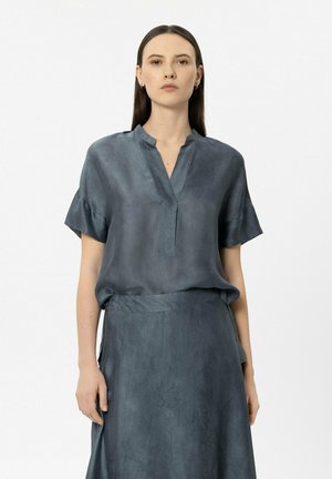 Frau mit langen, glatten Haaren, die eine locker sitzende dunkelgraue Kurzarmbluse und einen passenden Rock vor einem schlichten Hintergrund trägt.