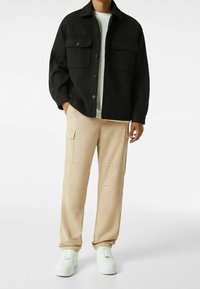 Svart mönstrad jacka med knäppning och två bröstfickor, i kombination med beige cargobyxor och vita sneakers. Avslappnad outfit.