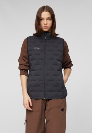 Junge Frau trägt eine schwarze gesteppte Weste über einem braunen Langarmshirt und braunen Hosen, steht mit einer Hand in der Tasche vor neutralem Hintergrund.
