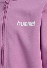 Gros plan d'une veste rose zippée avec un tissu texturé et le logo blanc "hummel" sur la poitrine gauche.
