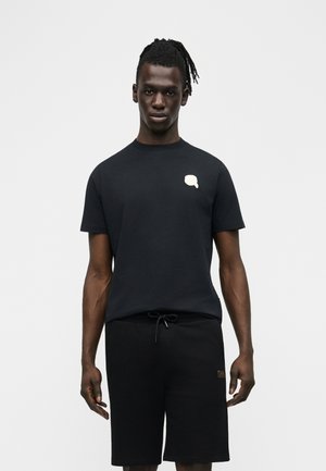 Homme aux dreadlocks portant un t-shirt noir avec un logo de bulle de dialogue blanche et un short noir à cordon, sur un fond blanc uni.