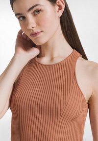 Rdzawoczerwony prążkowany top z wysokim kołnierzem i bez rękawów. Materiał wydaje się miękki, z pionową fakturą i bezszwowymi krawędziami.