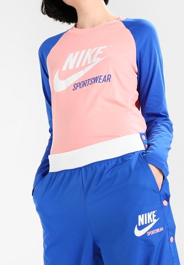 Mikina s raglánovými rukávy v růžové a modré barvě, se bílým logem Nike. Modré sportovní kalhoty s knoflíky a bílým pasem, také s brandingem Nike.