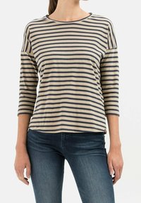 Woman wearing beige and navy striped long-sleeve shirt and dark blue jeans, standing against a plain white background.