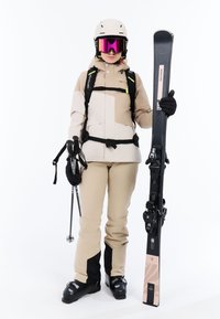 Schneesportausrüstung: beiger und schwarzer Skianzug, weißer Helm, pinke Skibrille, hält schwarze Skier und Skistöcke. Glattes Material, robuste Konstruktion.