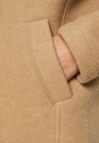 Detail eines braunen Wollmantels, der eine seitliche Tasche und weiches, strukturiertes Gewebe hervorhebt. Sichtbare Nähte und ein Teil einer Hand in der Tasche erkennbar.