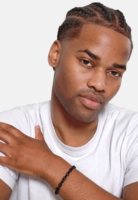 Black chain bracelet on a wrist, highlighting smooth links and a glossy finish. The wearer has short, braided hair and is in a white shirt.