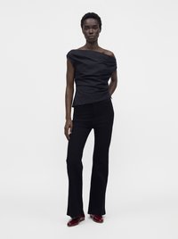 Femme debout avec assurance, portant un top noir épaules dénudées, un pantalon noir évasé et des chaussures rouge foncé sur un fond clair uni.