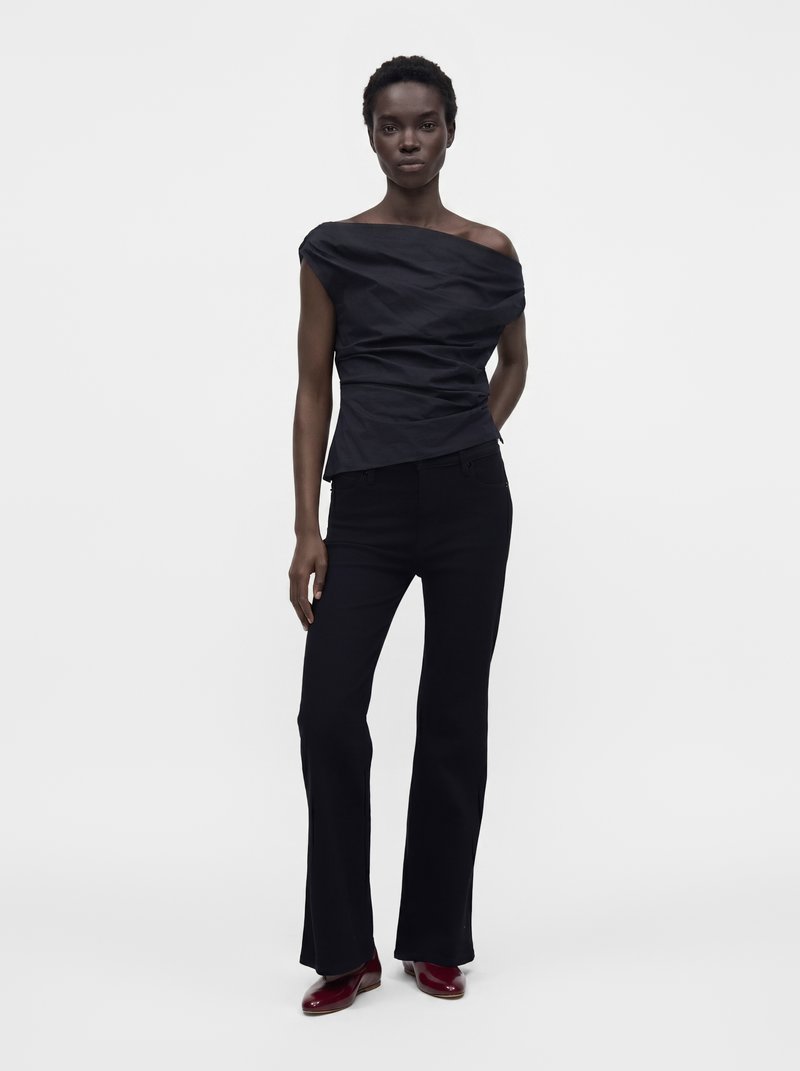 Woman stands confidently wearing a black off-shoulder top, black flared pants, and dark red shoes against a plain light background.