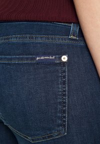 Los jeans de mezclilla azul oscuro presentan un bolsillo trasero con una etiqueta cosida que dice "para toda la humanidad" y un acento de botón metálico.