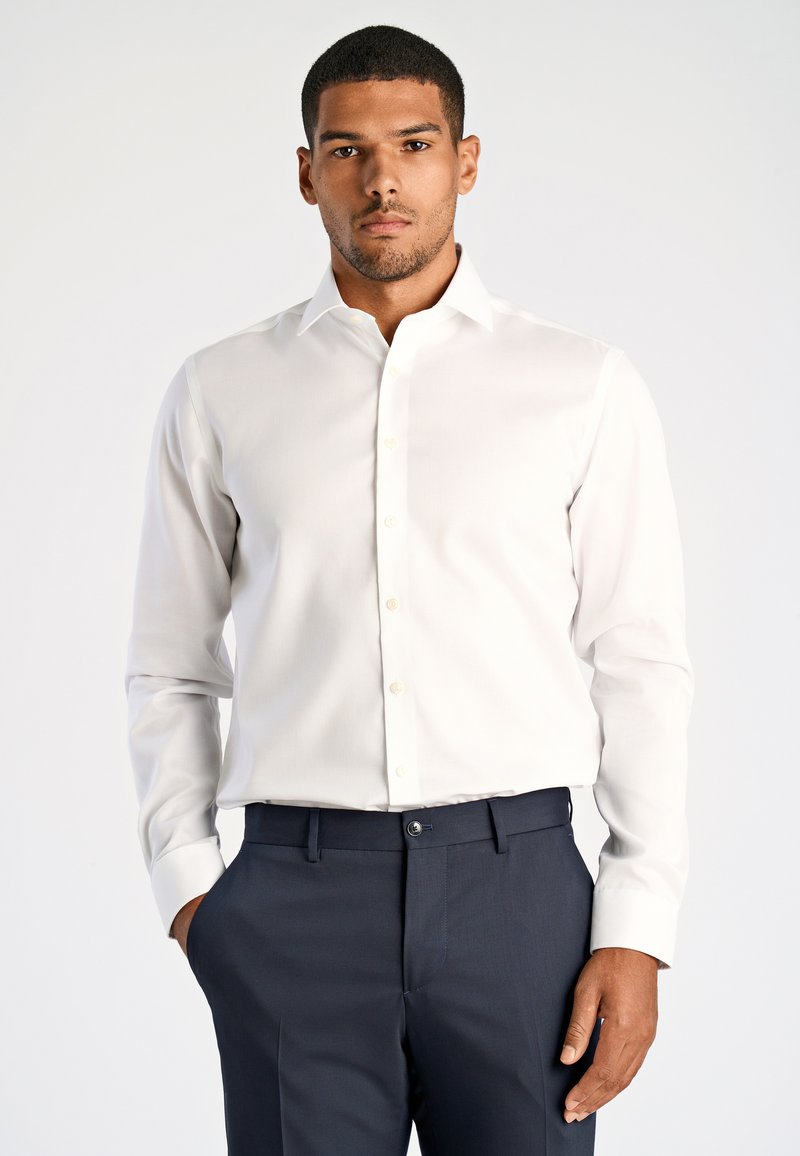 Camicia bianca con bottoni, collo classico, maniche lunghe e tessuto liscio, abbinata a pantaloni blu scuro.