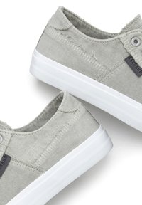 Hellgraue Leinen-Sneaker mit fransigen Kanten, weißen Gummisohlen und einem schwarzen Logo-Patch an der Seite.