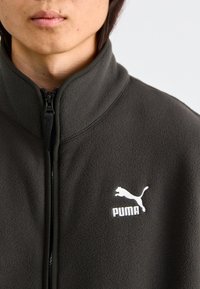 PUMA Fleecejacke, dunkelgrau, hoher Kragen, mit Reißverschluss vorne, weiche Textur, mit einem weißen Logo auf der linken Brustseite.