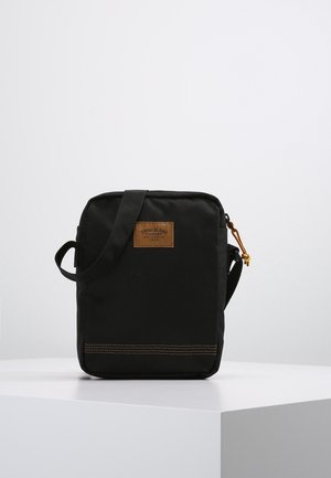 Czarna torba na ramię z płótna z przypinką ze skóry, zamknięciem na zamek i bocznym uchwytem. Posiada kontrastowe przeszycia u podstawy oraz beżowy akcent.