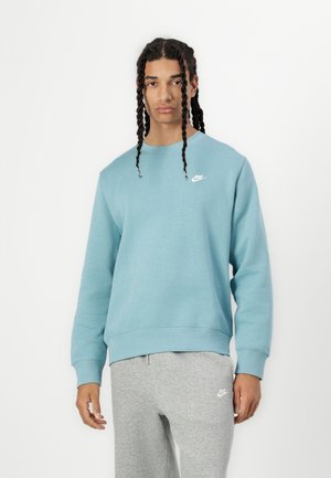 Sudadera Nike de color azul claro con cuello redondo, puños acanalados y un pequeño logo blanco en el lado izquierdo del pecho. Combinada con pantalones de chándal grises.