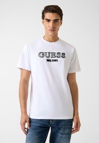 Biała bawełniana koszulka z nadrukiem "GUESS" i haftowanym napisem "EST. 1981" w czerni i szarości. Krótkie rękawy, okrągły dekolt, dopasowany krój.