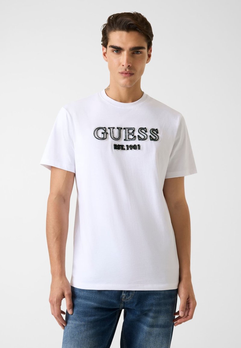 Biała bawełniana koszulka z nadrukiem "GUESS" i haftowanym napisem "EST. 1981" w czerni i szarości. Krótkie rękawy, okrągły dekolt, dopasowany krój.