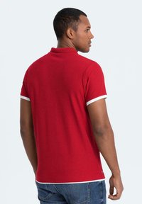 Röd polo med vita krageaccenter, korta ärmar, mjukt material och en skräddarsydd passform, visad bakifrån mot en enhetlig bakgrund.