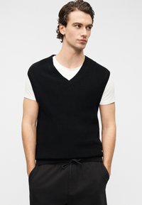 Μαύρο πλεκτό αμάνικο πουλόβερ με βαθύ V-λαιμό, φορεμένο πάνω από ένα λευκό T-shirt. Η υφή είναι ριμπ και έχει άνετη εφαρμογή.