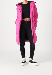 Abrigo acolchado rosa con capucha y cierre frontal, que cuenta con bolsillos laterales y una silueta larga. Se lleva sobre un top corto negro y jeans negros.