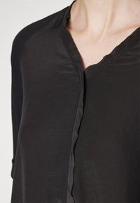 Personne portant un haut noir à manches longues avec un col en V et un détail de couture asymétrique sur le devant.