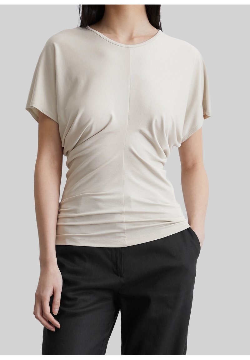 Femme portant un haut froncé beige à manches courtes rentré dans un pantalon noir, debout avec une main dans la poche sur fond gris.