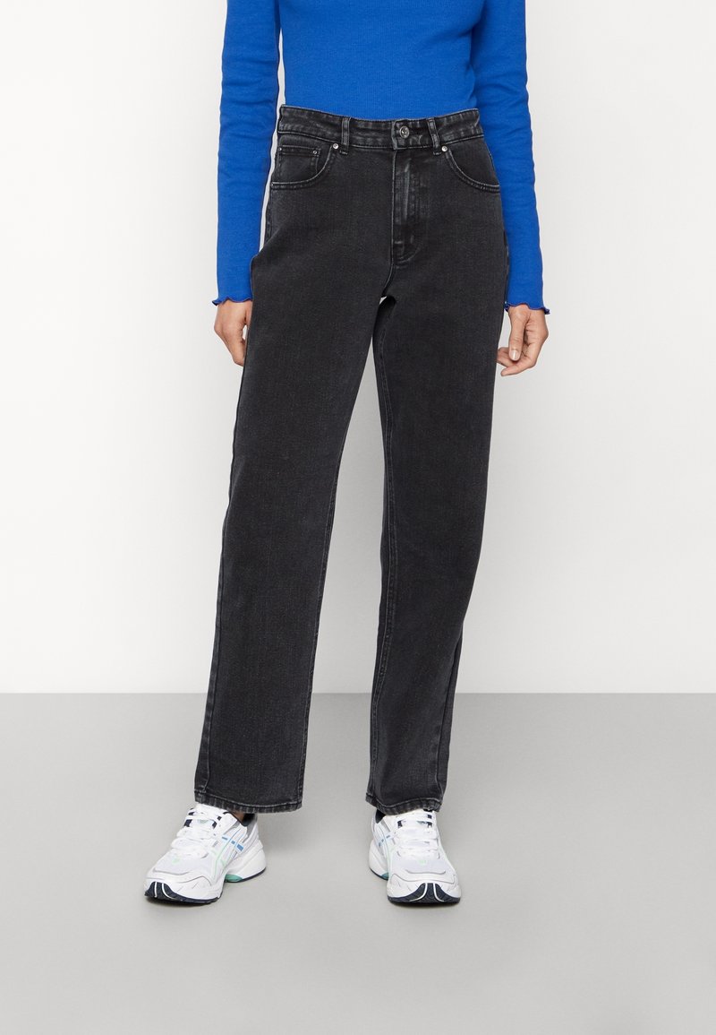 Personne portant un jean noir droit, un haut bleu vif à manches longues et des baskets blanches, debout sur un sol gris devant un mur blanc.