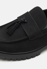 Sorte semskede loafers med flat såle, med en bred rem og dekorativ duskdetalj. Glatt tekstur og minimalistisk design.