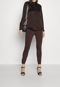 Ciemnobrązowa satynowa koszula z guzikami w połączeniu z brązowymi legginsami z skórzanymi akcentami. Modelka trzyma małą czarną torebkę na ramię i nosi czarne sandały na obcasie.