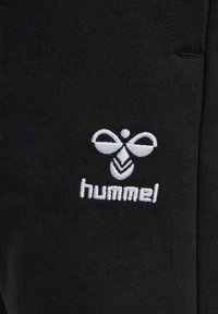Schwarze Jogginghose aus einem glatten Stoff. Enthält ein weißes, gesticktes Logo von "hummel" und ein bienenähnliches Emblem über dem Text.