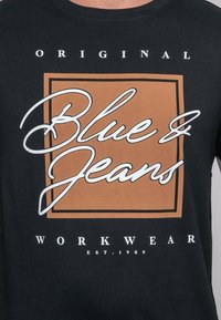 T-shirt en coton noir avec un motif central représentant le texte "Blue Jeans" en cursive blanche à l'intérieur d'un carré marron, étiqueté "WORKWEAR."