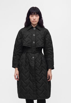 Femme portant un long manteau matelassé noir avec des boutons et une large ceinture, debout devant un fond clair uni.