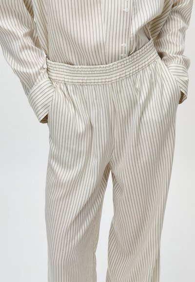 Άτομο που φοράει άνετο κρεμ παντελόνι και αντίστοιχο πουκάμισο με κουμπιά, με λεπτές κάθετες μαύρες ρίγες, με τα χέρια στις τσέπες.