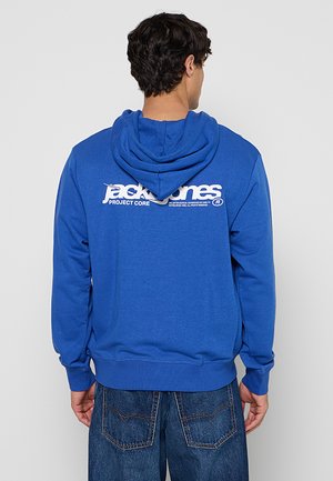 Man draagt een blauwe hoodie met een wit "jack & jones PROJECT CORE" logo op de rug, gecombineerd met blauwe spijkerbroek, met de rug naar de camera gekeerd.