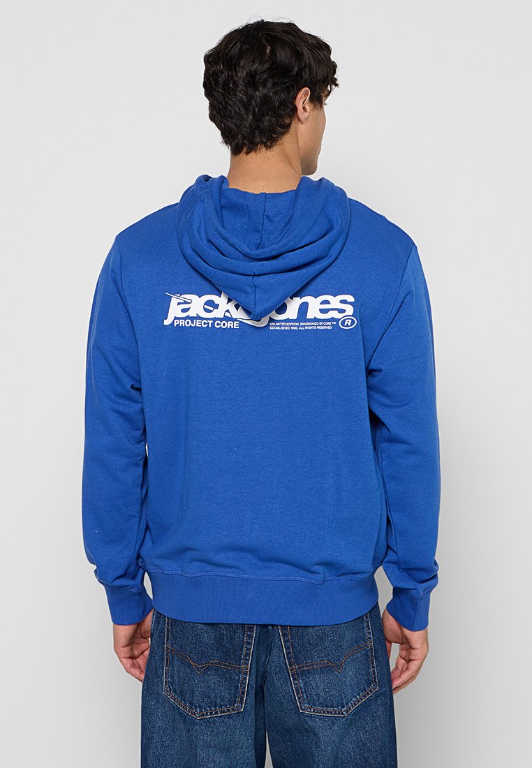 Άνδρας που φοράει μπλε φούτερ με κουκούλα με λευκό λογότυπο "jack & jones PROJECT CORE" στην πλάτη, σε συνδυασμό με μπλε τζιν, με την πλάτη προς την κάμερα.
