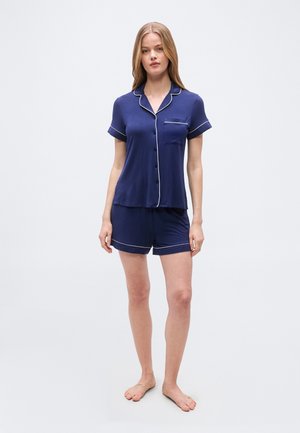 Vrouw staat blootsvoets en draagt een marineblauw korte mouwen pyjamashirt met knopen en een bijpassende korte broek met witte biezen aan de mouwen en randen.
