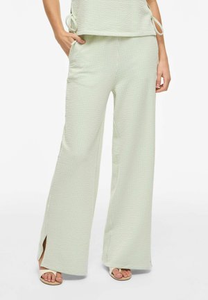 Kobieta ubrana w jasnozielone, teksturowane spodnie o szerokich nogawkach z bocznymi rozcięciami, zestawione z pasującym topem i beżowymi sandałami na otwartym palcu.