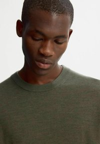 Kaki-grön t-shirt med rund halsringning och korta ärmar, tillverkad av mjukt, texturerat tyg; synlig sömnad runt halsringningen.