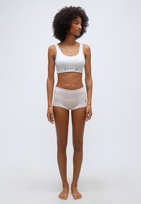 Λεπτή γυναίκα με αφρο μαλλιά, όρθια ξυπόλητη, φορώντας λευκό αθλητικό μπουστάκι Polo και αντίστοιχα boxer σορτς μπροστά σε απλό ανοιχτό φόντο.