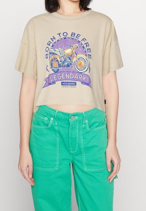 Beige cropped T-shirt med lilla og orange vintage motorcykelmotiv, tekst "Born to Be Free Legendary Wild Riders," båret med grønne bukser.