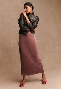 Schwarze Lederjacke mit Reißverschlussdetails, kombiniert mit einem langen, roséfarbenen Satten Rock und rosa Absatzschuhen mit Schleifenakzenten, was einen geschichteten Look erzeugt.